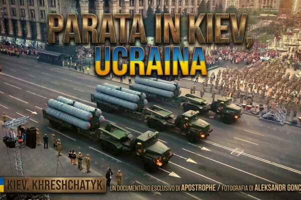 Una veduta aerea di una parata militare che si svolge su Via Khreshchatyk a Kiev, in Ucraina. La strada è percorsa da tre enormi camion trasportatori che esibiscono fieri un totale di nove missili a lungo raggio. Al di là del corteo, si distinguono chiaramente edifici storici monumentali e una folla di spettatori che assiste all'evento. Un testo sovrapposto in grande carattere e in alto recita: "PARATA IN KIEV, UCRAINA". In basso a sinistra, una bandiera ucraina affianca la dicitura "KIEV, KHRESHCHATYK". In basso a destra, sono indicati i crediti: "UN DOCUMENTARIO ESCLUSIVO DI APOSTROPHE / FOTOGRAFIA DI OLEKSANDR HONCHAROV